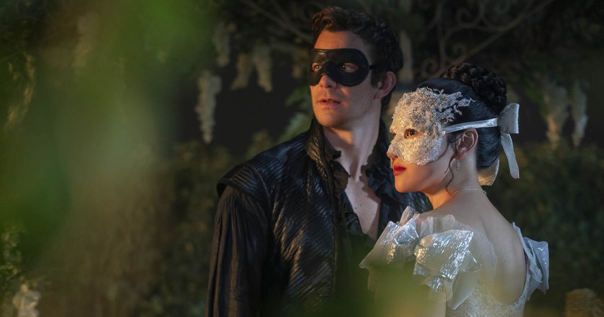 A couple wearing masks at an elegant outdoor setting, with the man in a black costume and the woman in a white dress adorned with a ribbon, surrounded by lush greenery.